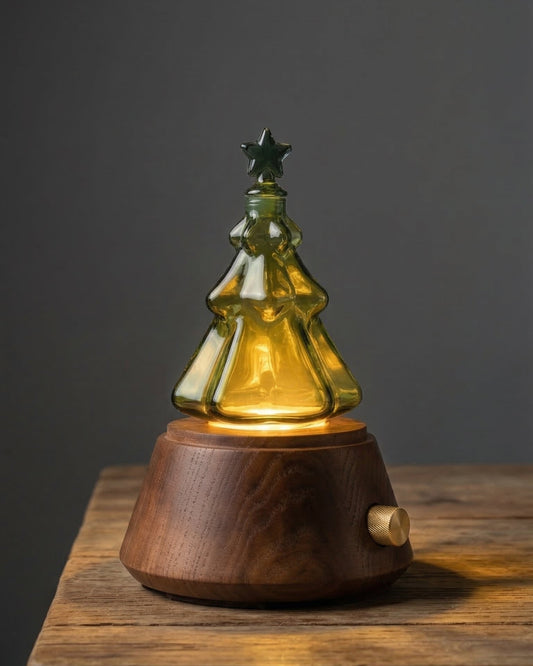 A professional studio product photograph of a Charabanc nebulizing aromatherapy diffuser. The device features a multi-tiered clear glass flask expertly shaped like a Christmas tree with a star on top. It sits on a premium round dark walnut wood base with a signature knurled gold control knob. The image is set against a dark, solid background to highlight the elegant, festive design and high-quality craftsmanship of the waterless essential oil diffuser.