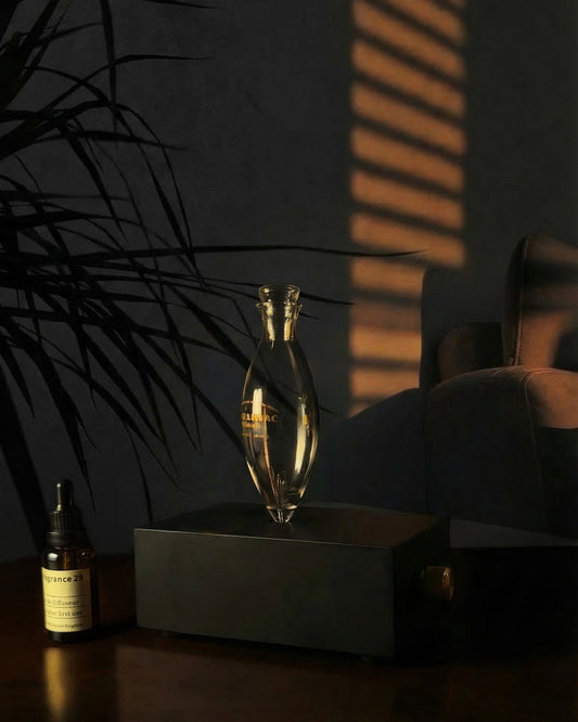 A sleek black rectangular Charabanc Travel Mini nebulizing essential oil diffuser, featuring a clear glass nebula flask and a knurled gold control knob. This portable cordless rechargeable device is shown on a dark wooden table next to an amber essential oil bottle. Warm sunset light filters through window blinds, casting a dramatic shadow of a houseplant on the wall, creating a moody, luxury atmosphere.