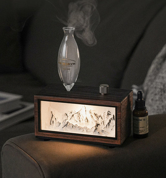 "A serene lifestyle shot of the Charabanc 'Alpine Edition' waterless nebulizer. The device features a sleek white console housing a detailed 3D mountain relief, illuminated by a warm golden light that mimics the alpenglow. Placed on a sofa next to a person reading, creating a perfect Zen meditation atmosphere."