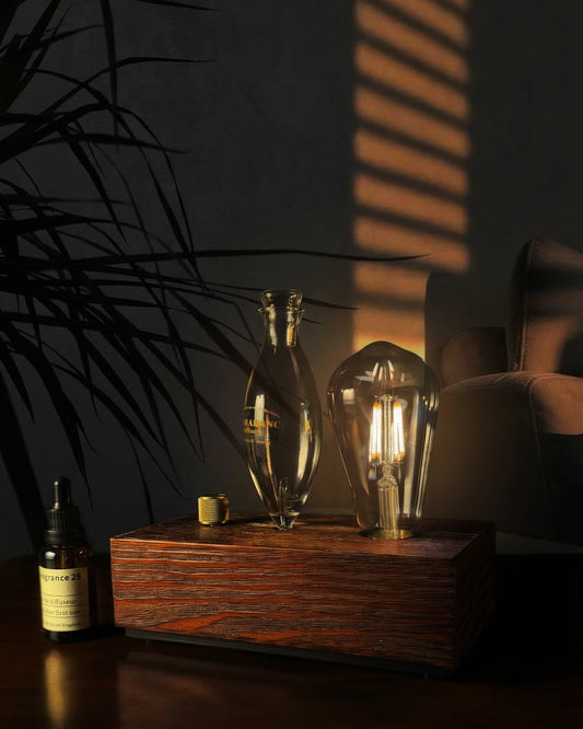 "A Charabanc Fragrance waterless nebulizing diffuser featuring an illuminated Edison-style bulb that casts a warm amber glow. The instrument is crafted with a hand-blown glass nebulizer and a solid dark wood base, elegantly placed on a wooden table next to a signature amber essential oil bottle in a moody, sophisticated interior."