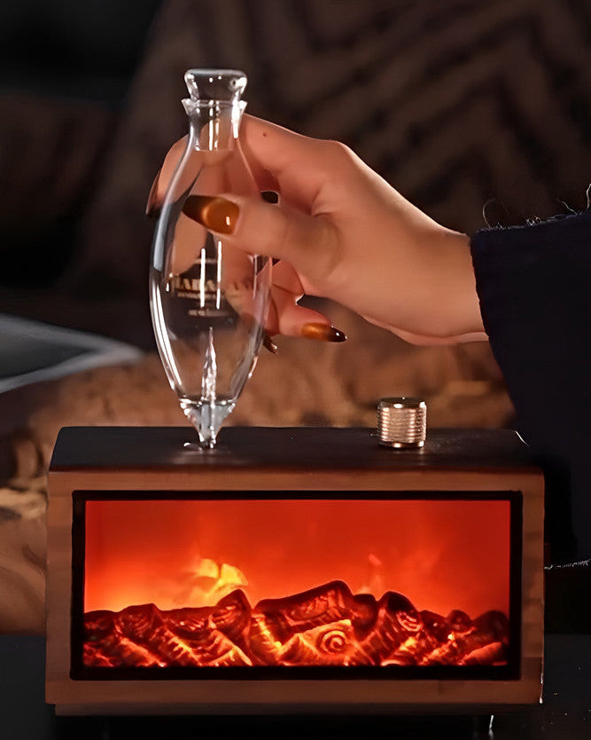 "A lifestyle photograph showing hands gently touching the dark wood console of the Charabanc 'Hearth Edition' waterless nebulizer. The realistic fireplace flame effect is glowing warmly inside the base, and steam is actively rising from the hand-blown glass chamber."