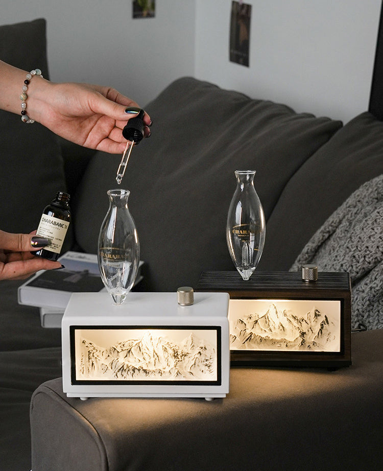 "A serene lifestyle shot of the Charabanc 'Alpine Edition' waterless nebulizer. The device features a sleek white console housing a detailed 3D mountain relief, illuminated by a warm golden light that mimics the alpenglow. Placed on a sofa next to a person reading, creating a perfect Zen meditation atmosphere."