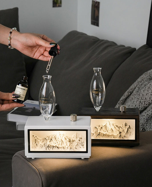 "A serene lifestyle shot of the Charabanc 'Alpine Edition' waterless nebulizer. The device features a sleek white console housing a detailed 3D mountain relief, illuminated by a warm golden light that mimics the alpenglow. Placed on a sofa next to a person reading, creating a perfect Zen meditation atmosphere."