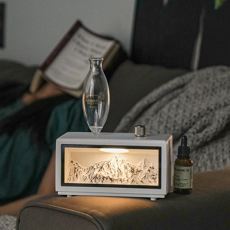 "A serene lifestyle shot of the Charabanc 'Alpine Edition' waterless nebulizer. The device features a sleek white console housing a detailed 3D mountain relief, illuminated by a warm golden light that mimics the alpenglow. Placed on a sofa next to a person reading, creating a perfect Zen meditation atmosphere."