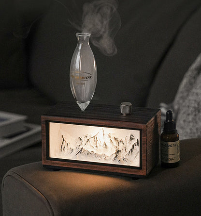 "A serene lifestyle shot of the Charabanc 'Alpine Edition' waterless nebulizer. The device features a sleek white console housing a detailed 3D mountain relief, illuminated by a warm golden light that mimics the alpenglow. Placed on a sofa next to a person reading, creating a perfect Zen meditation atmosphere."