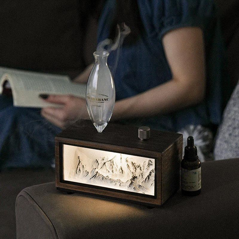 "A serene lifestyle shot of the Charabanc 'Alpine Edition' waterless nebulizer. The device features a sleek white console housing a detailed 3D mountain relief, illuminated by a warm golden light that mimics the alpenglow. Placed on a sofa next to a person reading, creating a perfect Zen meditation atmosphere."
