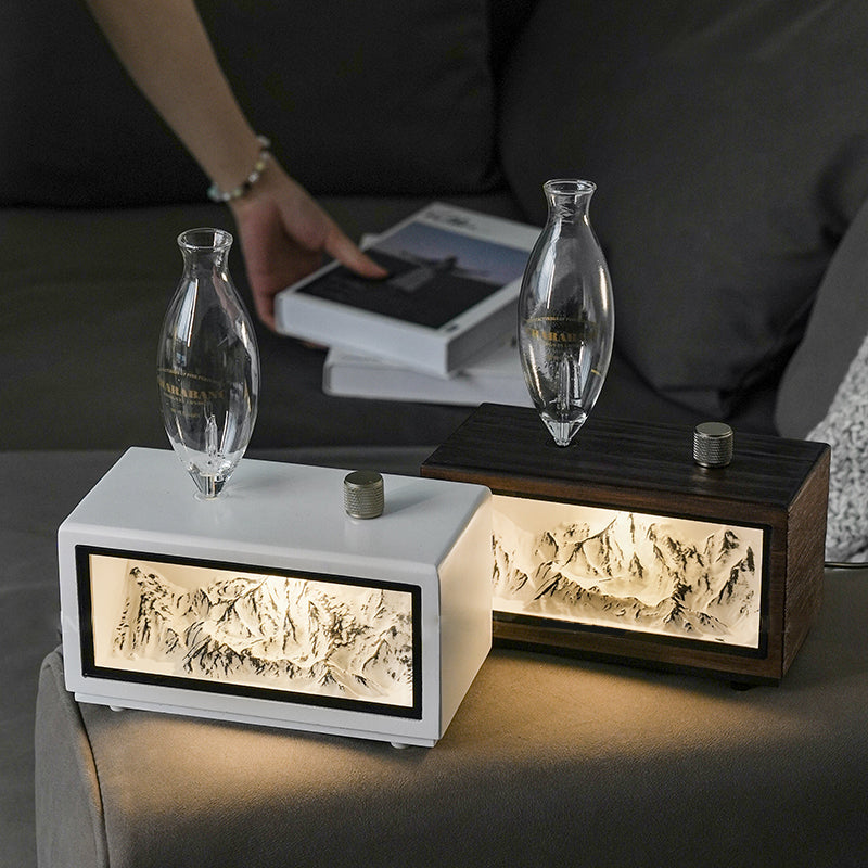 "A serene lifestyle shot of the Charabanc 'Alpine Edition' waterless nebulizer. The device features a sleek white console housing a detailed 3D mountain relief, illuminated by a warm golden light that mimics the alpenglow. Placed on a sofa next to a person reading, creating a perfect Zen meditation atmosphere."