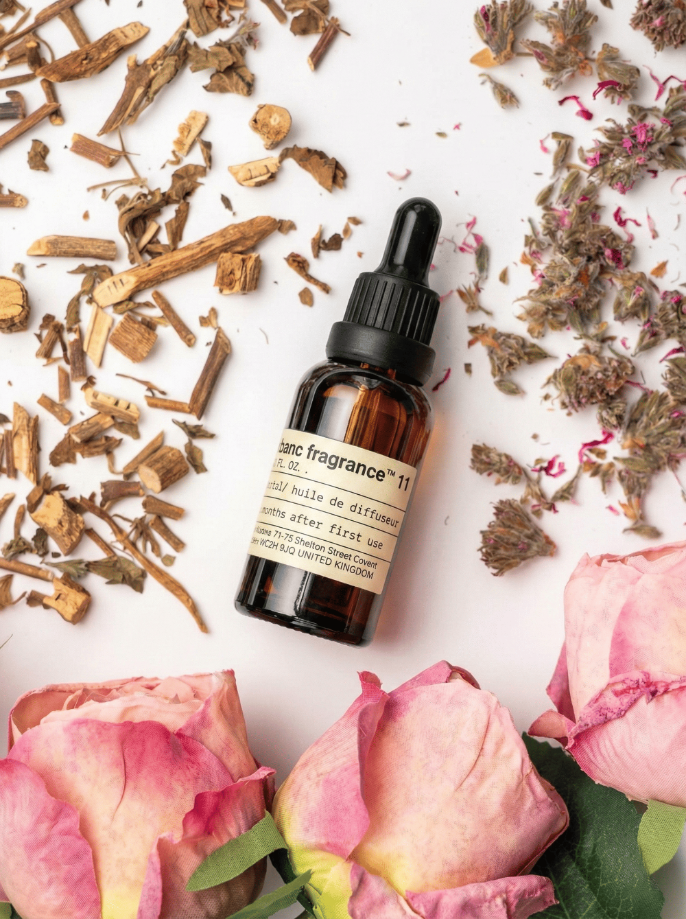 Charabanc Fragrance NO.11 Rose Whisper essential oil bottle surrounded by blooming pink roses, dried rosebuds, and earthy botanical accents on a white surface.