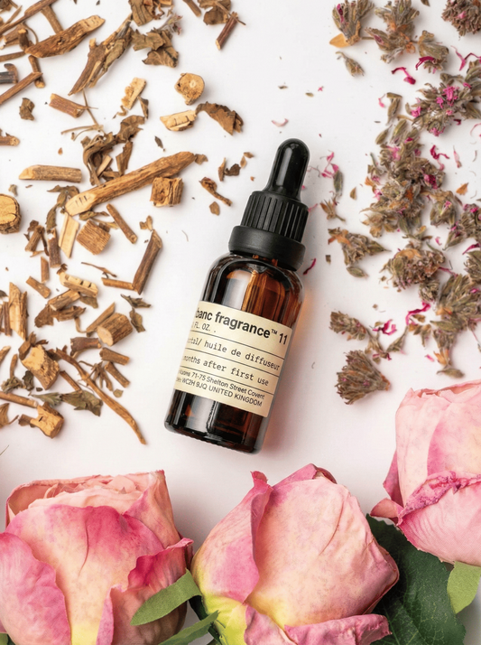 Charabanc Fragrance NO.11 Rose Whisper essential oil bottle surrounded by blooming pink roses, dried rosebuds, and earthy botanical accents on a white surface.