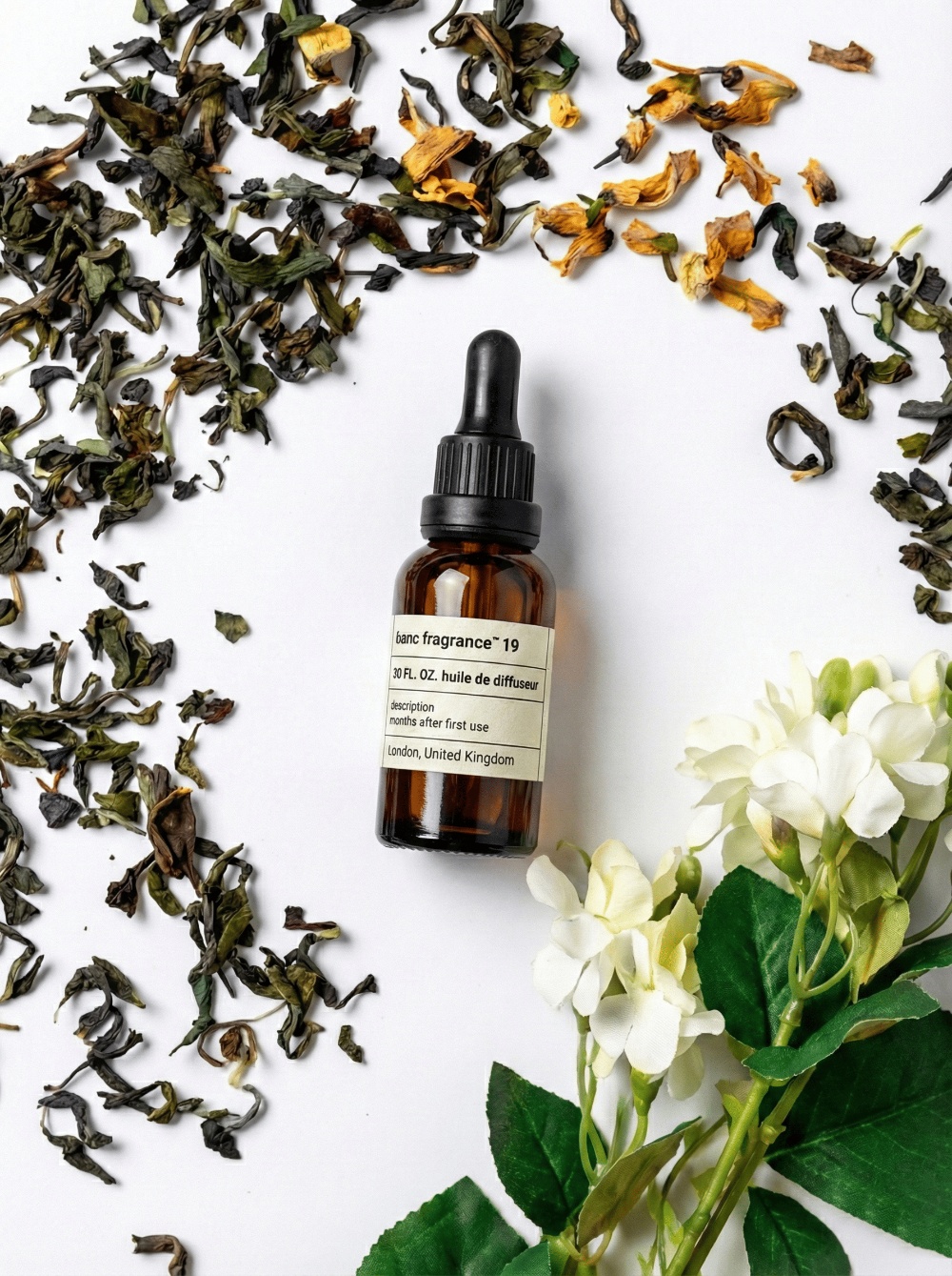 Charabanc Fragrance NO.19 All-Season Zen Tea essential oil bottle surrounded by loose green tea leaves and white jasmine flowers, evoking a serene tea ritual.