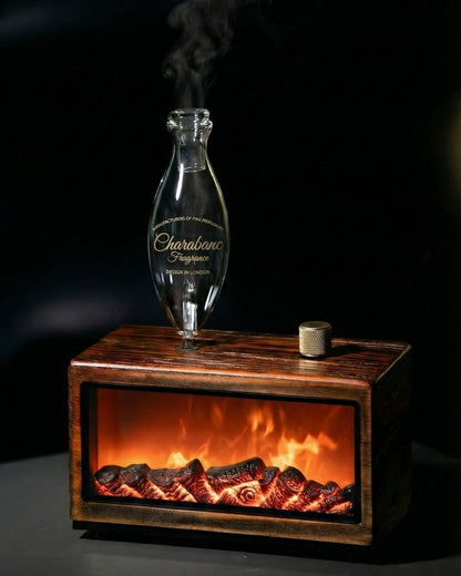"A lifestyle photograph showing hands gently touching the dark wood console of the Charabanc 'Hearth Edition' waterless nebulizer. The realistic fireplace flame effect is glowing warmly inside the base, and steam is actively rising from the hand-blown glass chamber."