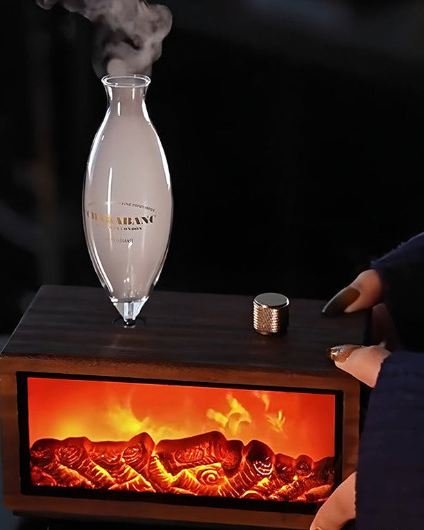 "A lifestyle photograph showing hands gently touching the dark wood console of the Charabanc 'Hearth Edition' waterless nebulizer. The realistic fireplace flame effect is glowing warmly inside the base, and steam is actively rising from the hand-blown glass chamber."
