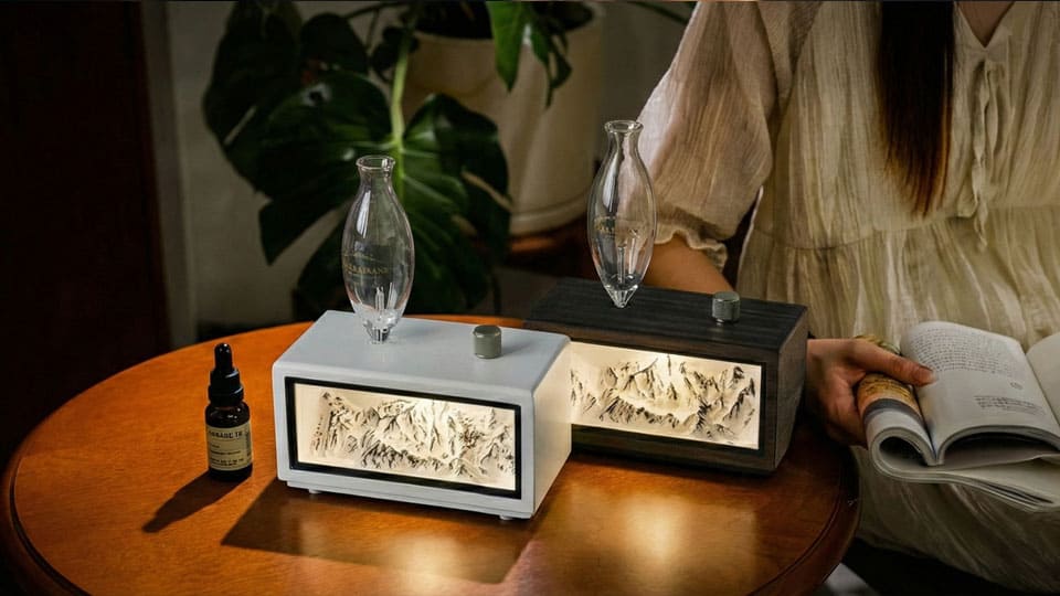 "Two Charabanc waterless essential oil diffusers, one in white and one in dark wood, featuring an illuminated miniature mountain range carved into the front panel. The diffusers sit on a wooden table next to a bottle of oil, while a woman sits in the background reading a book, creating a serene, contemplative atmosphere where light, landscape, and scent converge."