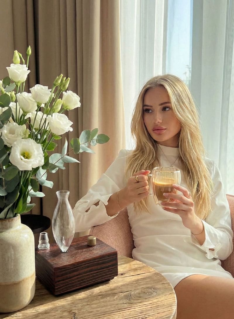 "A lifestyle photograph of a blonde woman in a white dress enjoying a cup of coffee in a sunlit room. On the rustic wooden side table next to her sits a Charabanc waterless diffuser and a vase of fresh white flowers, creating an atmosphere of quiet elegance and sensory indulgence."