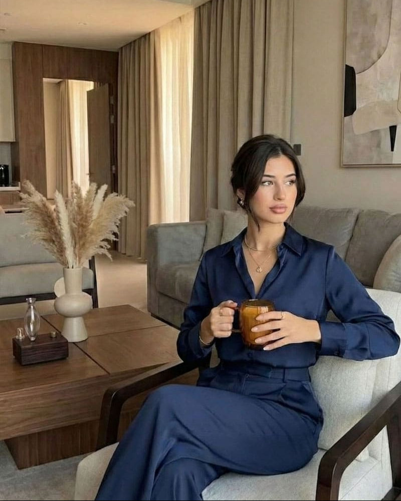 "A sophisticated lifestyle shot in a high-end modern apartment. A woman in a navy blue silk blouse and matching trousers sits elegantly on a light grey armchair, holding a glass of coffee. In the background, the Charabanc waterless scent diffuser with a walnut wood base is placed on a dark wood coffee table next to a minimalist pampas grass arrangement, blending professional style with domestic serenity."